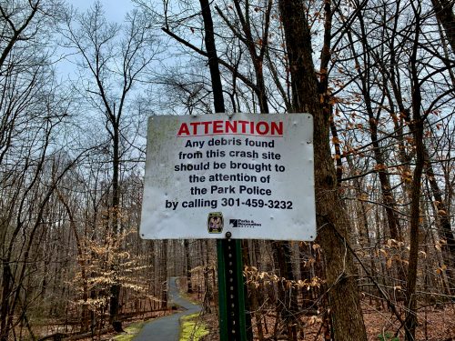 Sign in a wooded area instructing to report debris from a crash site to Park Police at 301-459-3232.