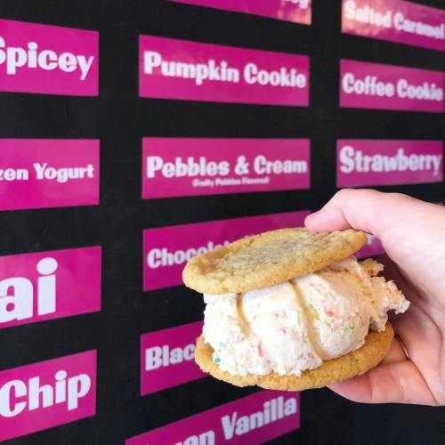 A hand holds a cookie sandwich filled with colorful ice cream, with a menu of flavors in the background.