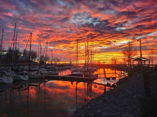 A vibrant sunset over a marina, with colorful clouds reflecting on the water and silhouetted sailboats.