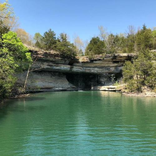 A serene natural landscape featuring a calm green lake surrounded by lush trees and rocky cliffs.