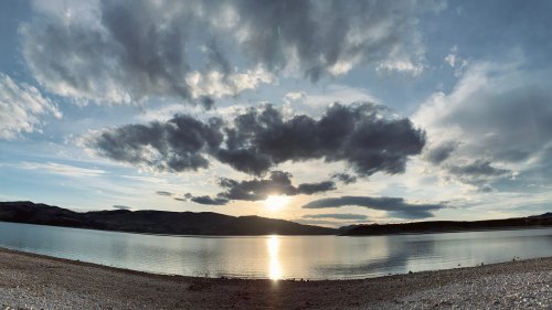 A serene lakeside view at sunset, with clouds reflecting on the water and mountains in the background.