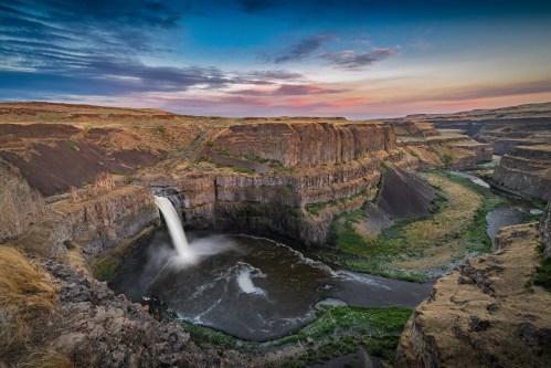 A stunning waterfall cascades into a river, surrounded by rugged cliffs and a colorful sunset sky.