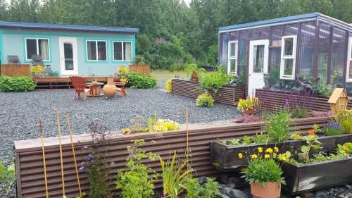 A colorful garden with flower beds, two buildings, and a greenhouse surrounded by gravel and greenery.