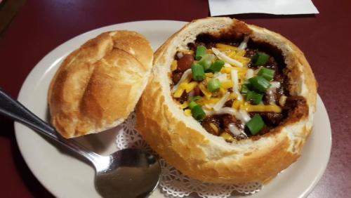 A bread bowl filled with chili, topped with cheese and green onions, served with a side of bread.
