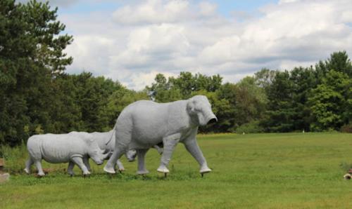Two large, gray animal sculptures resembling an elephant and a rhino walking on a grassy field under a cloudy sky.