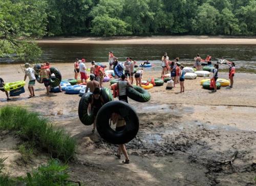 People carrying inner tubes at a riverbank, preparing for a day of tubing in a lush, green environment.