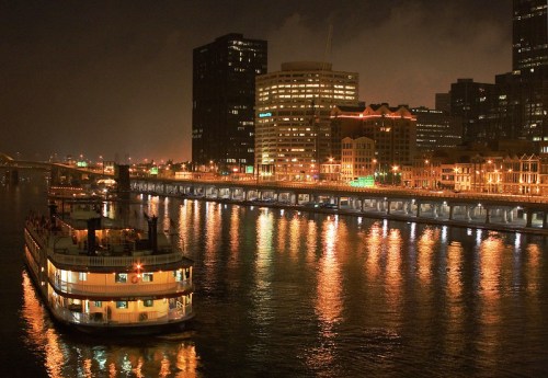 A nighttime cityscape with a boat on a river, reflecting lights from buildings along the shore.