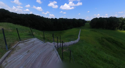 A wooden pathway winds down a grassy hill, surrounded by trees under a bright blue sky with fluffy clouds.
