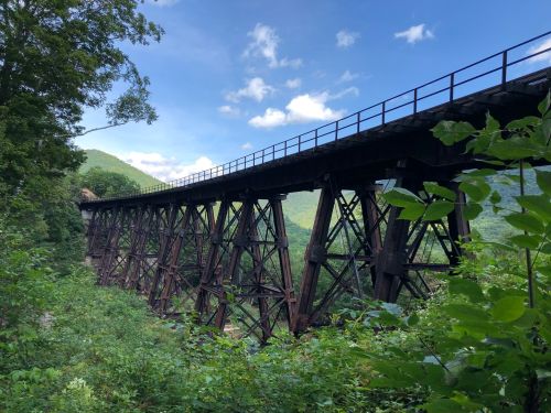 A tall, rusted railway bridge spans a valley, surrounded by lush green trees and rolling hills under a blue sky.