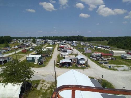 Aerial view of a rural market with various stalls and vehicles, surrounded by greenery and blue skies.