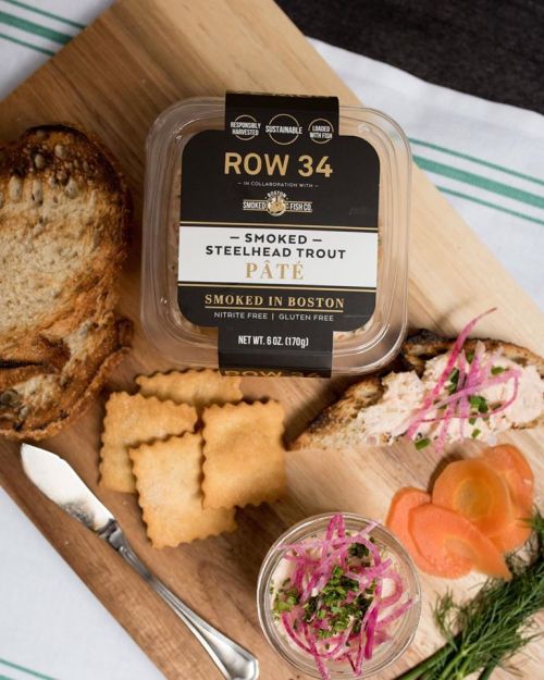A wooden board with smoked steelhead trout pâté, crackers, bread, pickled vegetables, and a fork.