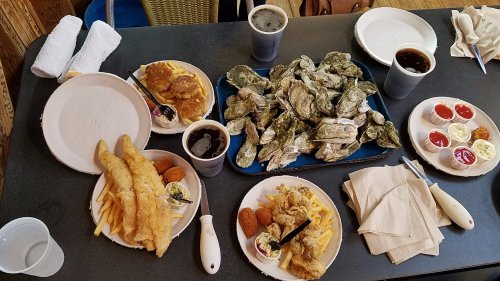A table spread with plates of fried fish, oysters, fries, and drinks, accompanied by dipping sauces.