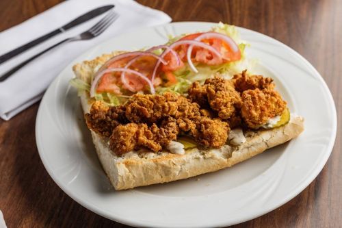 A plate with a sandwich featuring fried chicken, lettuce, tomatoes, pickles, and mayonnaise on toasted bread.