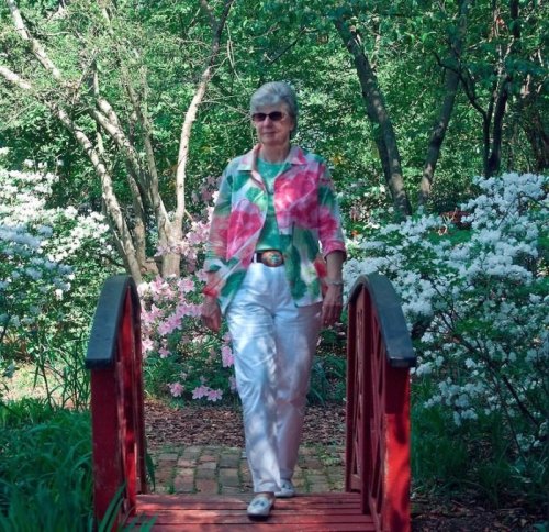 A woman in a colorful floral shirt walks across a red bridge surrounded by blooming azaleas in a lush garden.