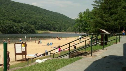 A sandy beach by a lake surrounded by green hills, with people sunbathing and swimming on a sunny day.