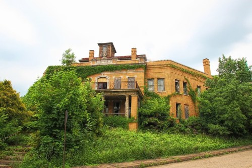 An overgrown, abandoned brick building surrounded by lush greenery and trees under a cloudy sky.