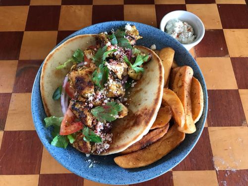 A plate of grilled chicken with vegetables and herbs, served with pita bread and crispy potato wedges, alongside a dip.