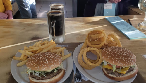 Two burgers with lettuce, cheese, and toppings, served with fries and onion rings, alongside two glasses of soda.