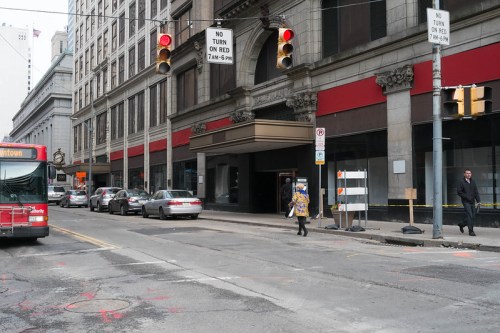 A city street with traffic lights, parked cars, a bus, and a person walking past a building with red awnings.