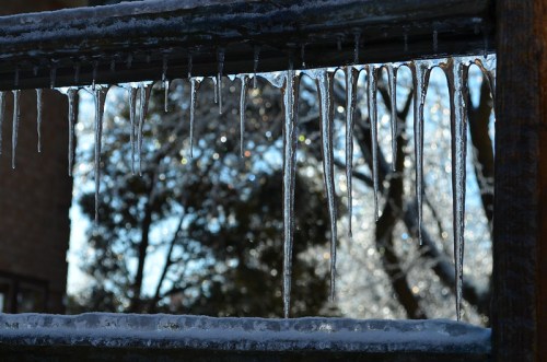 Icicles hang from a wooden railing, glistening in the sunlight with a blurred, sparkling background of trees.
