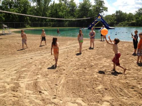 Children playing beach volleyball near a lake, with a water slide and trees in the background on a sunny day.