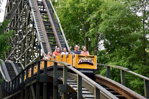 A roller coaster named "Phoenix" with excited riders waving as it climbs a steep track surrounded by trees.