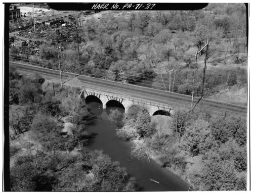 Aerial view of a stone arch bridge over a river, surrounded by trees and railway tracks.
