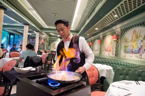 A chef in a formal outfit cooks at a restaurant, flames rising from a pan on a table in a stylish dining area.