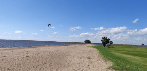 A sandy beach beside a calm lake, with green grass and a few trees under a blue sky with scattered clouds.