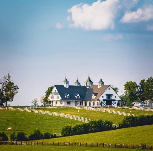 A picturesque farmhouse with three spires, surrounded by green fields and a white fence under a clear blue sky.