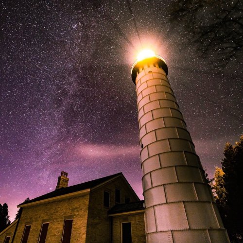 A tall lighthouse illuminated at night, surrounded by a starry sky and a faint glimpse of the Milky Way.