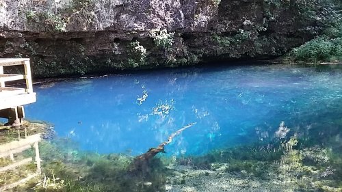 A serene blue spring surrounded by lush greenery and rocky cliffs, with a wooden platform nearby.