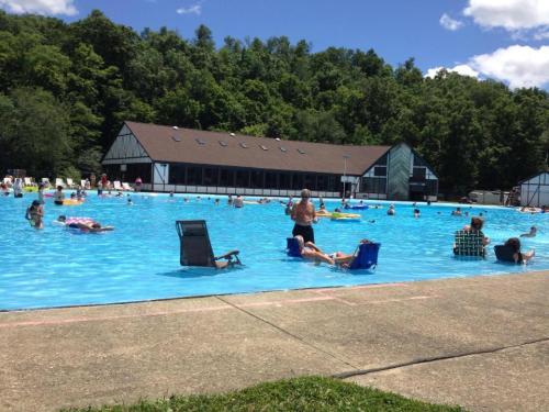 A large outdoor pool filled with people enjoying the sun, surrounded by trees and a building in the background.
