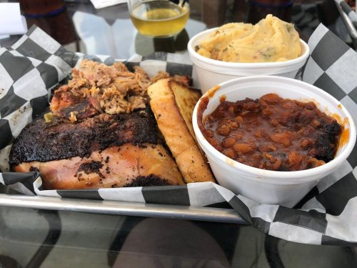 A tray with smoked meat, mashed potatoes, and baked beans, served with a slice of bread on a checkered paper.