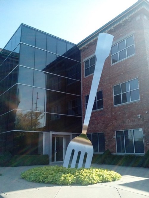 Giant metallic fork sculpture in front of a modern glass and brick building, surrounded by green landscaping.