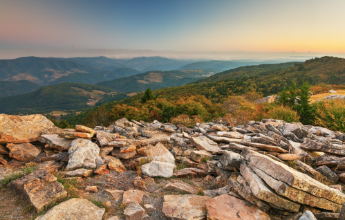 Scenic view of mountains and valleys at sunset, with rocky foreground and colorful autumn foliage.