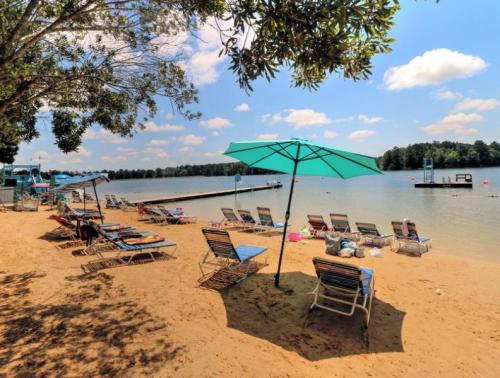 A sandy beach with lounge chairs and umbrellas, overlooking a calm lake under a bright blue sky.