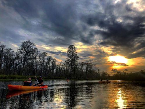 Two people kayak on a calm river at sunset, surrounded by trees and dramatic clouds reflecting in the water.