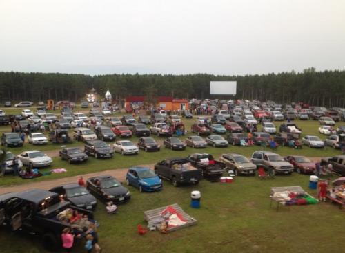 A crowded drive-in movie theater with cars parked in a grassy area and a large screen in the background.