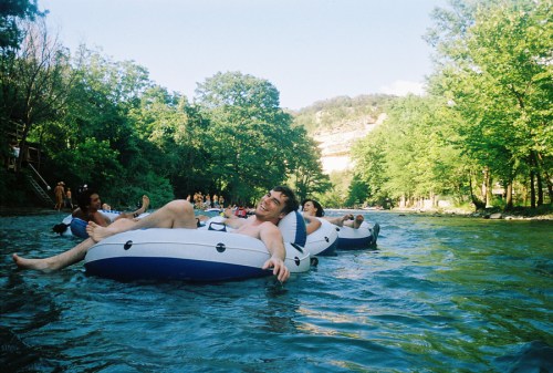 Three people relax on inflatable tubes in a river, surrounded by lush green trees and a sunny sky.