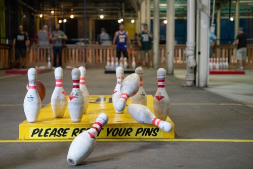 A bowling alley scene with pins scattered on the lane and players in the background preparing to bowl.
