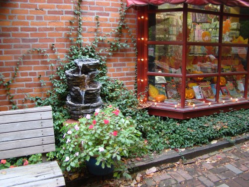 A cozy corner with a brick wall, a small fountain, flowers, and a window display featuring pumpkins and autumn decor.