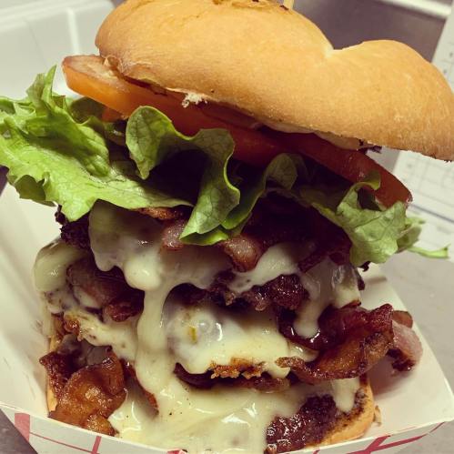 A towering burger with layers of bacon, melted cheese, lettuce, and tomato, served in a paper tray.