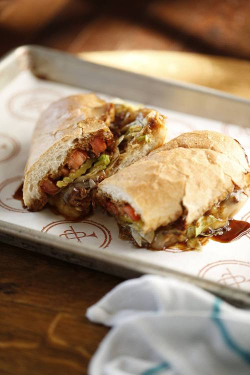 A close-up of a halved sandwich filled with meat, pickles, and vegetables, served on a tray with a logo.