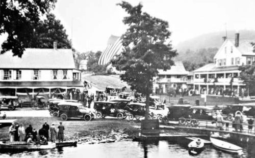 Historic black-and-white photo of a lakeside scene with vintage cars, buildings, and people gathered by the water.