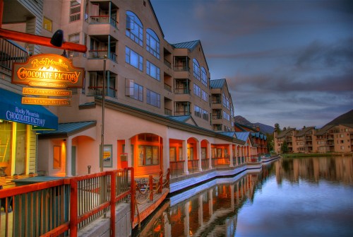 A scenic view of the Rocky Mountain Chocolate Factory by a calm lake, with buildings reflecting in the water at dusk.