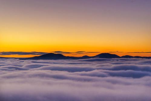 A serene landscape of mountains above a sea of clouds at sunrise, with a gradient of orange and purple in the sky.