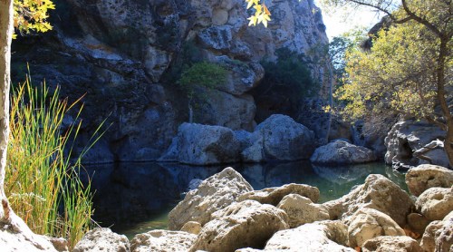 A serene rocky landscape with a calm pool of water surrounded by trees and cliffs under bright sunlight.
