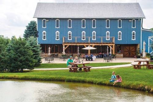 A blue barn with people sitting outside near a pond, surrounded by greenery and picnic tables.