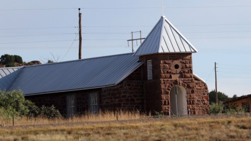 A stone church with a metal roof, surrounded by dry grass and power lines in the background.